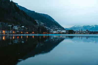 A town with glowing streetlights along a lake reflecting mountains under a cool sky, Ultra