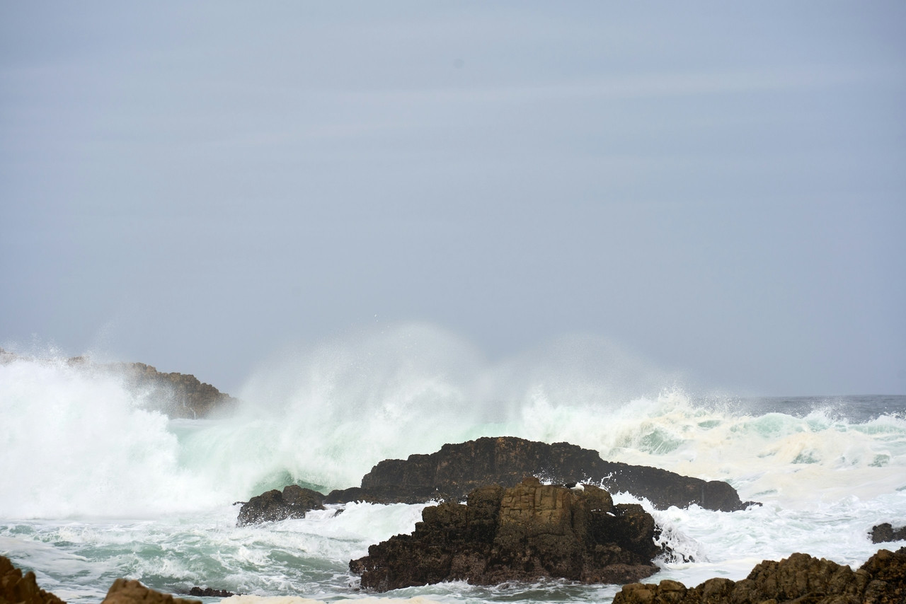 Large waves crash against dark rocks under a grey sky, Ultra