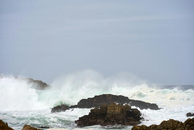 Large waves crash against dark rocks under a grey sky, Ultra